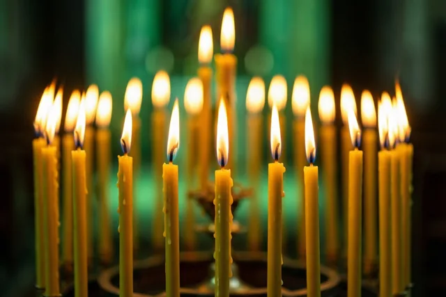 Een rij brandende gele kaarsen met smeltend kaarsvet staat in een donkere kerk tegen een onscherpe groene achtergrond.