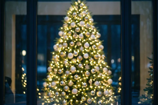 Door een donker raamkozijn is een onscherpe kerstboom te zien met gele en witte lichtjes tegen koele blauwe nachttinten.