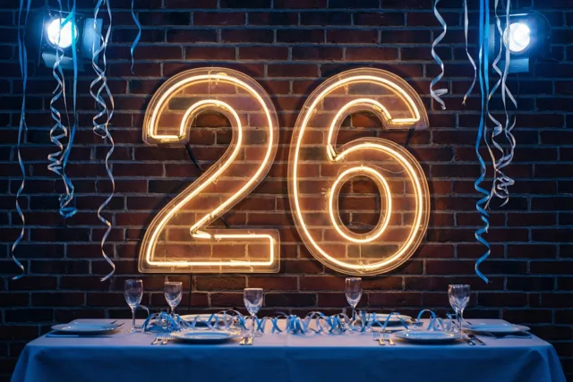Een geel neongetal 26 hangt aan een donkere bakstenen muur boven een tafel met borden, glazen en blauwe slingers, onder blauwe spots en hangende blauwe en zilveren slingers.