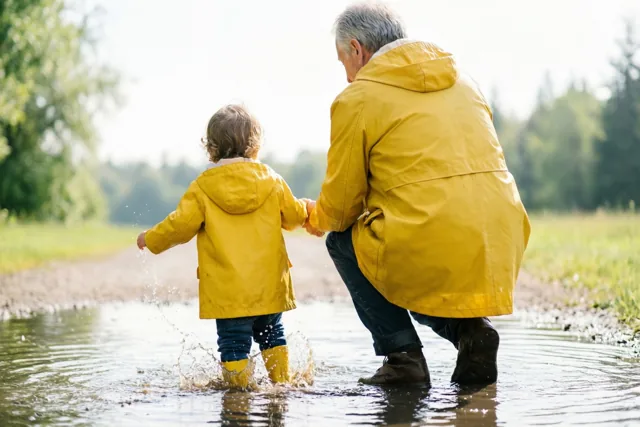 Een grootouder en een peuter stappen hand in hand door een ondiepe plas in dezelfde felgele regenjassen en gele regenlaarzen, van achteren gezien met opspattend water.