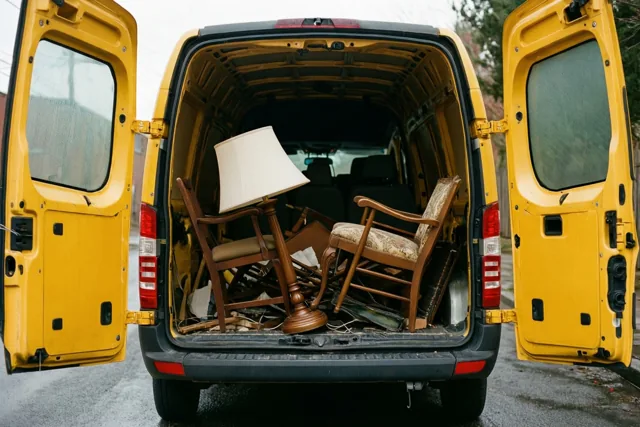 Een gele verhuiswagen staat op een natte verharding onder een bewolkte hemel, met open achterdeuren en uitstekende meubels, waaronder een stoel en een lamp.