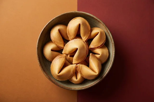 Een keramieken kom met goudbruine gelukskoekjes staat op een ondergrond met een oranje en een dieprood vlak.