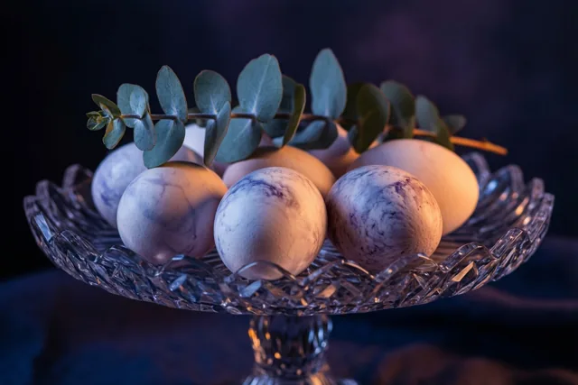 Een kristallen taartplateau met lichtgekleurde gemarmerde eieren en een takje eucalyptus staat tegen een donkere achtergrond, belicht met warme en koele tinten.