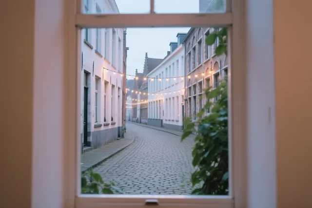 Door een raamkozijn is een Gentse kasseistraat te zien met oude gebouwen en warme lichtslingers ertussen, in zacht koel licht.