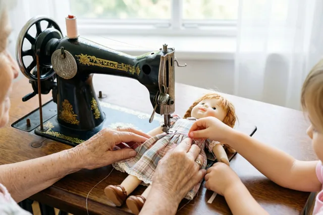 Een oudere vrouw en een jong kind naaien een geruit jurkje aan een pop bij een oude zwarte naaimachine op een houten tafel naast het raam.