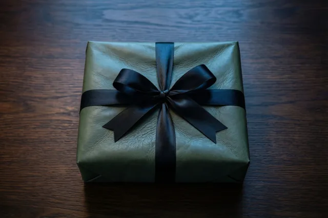 Een geschenk in olijfgroen leer met een zwarte satijnen strik ligt op een donkere eiken tafel tussen diepe schaduwen.