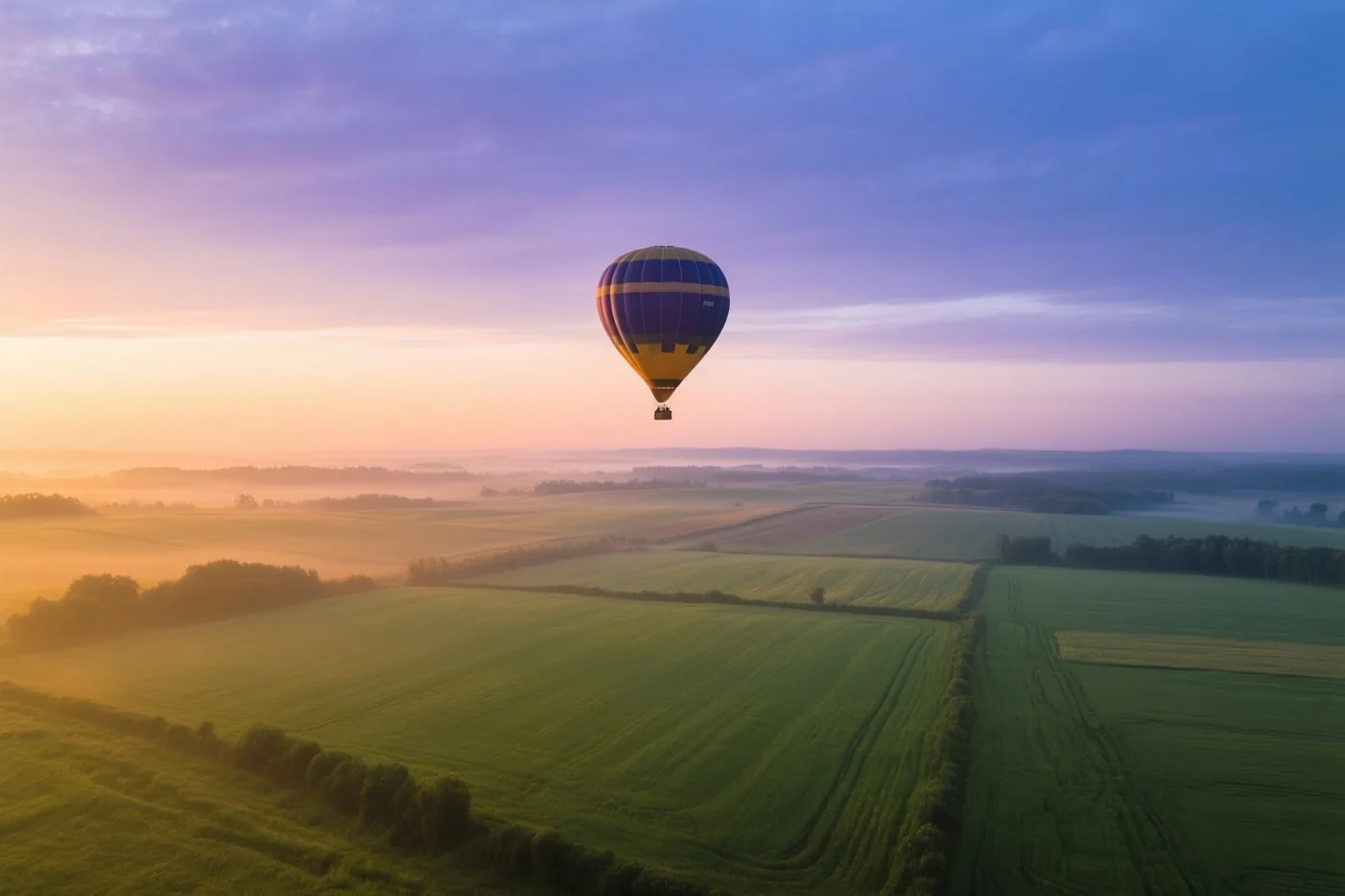 Een blauw-geel gestreepte luchtballon zweeft bij zonsopgang boven groene velden onder een paarse, roze en oranje lucht.