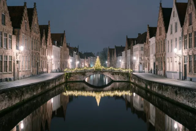 Historische gebouwen en een brug met kerstverlichting weerspiegelen in een waterloop onder de donkerblauwe nachthemel van een oud Vlaams stadje.
