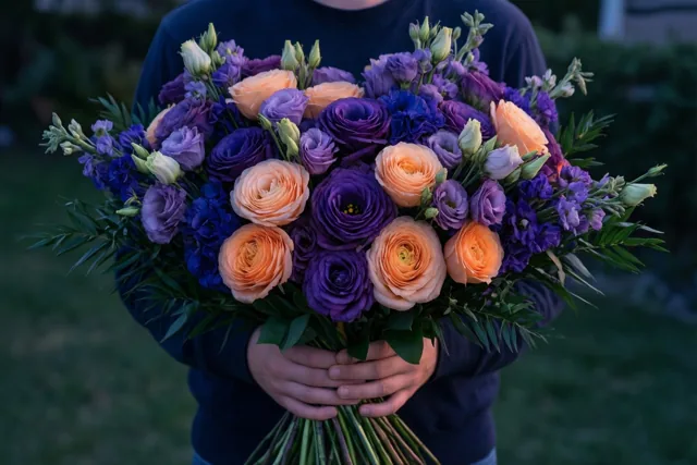 Iemand in een donker shirt houdt buiten in gedempt daglicht een groot boeket paarse en perzikkleurige bloemen vast dat het gezicht verbergt.