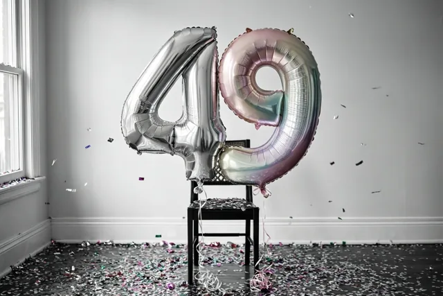 Twee grote folieballonnen in de vorm van 4 en 9, zilver en met een parelmoerglans in pasteltinten, zijn vastgemaakt aan een zwarte stoel in een lichte kamer met confetti, een witte muur en een raam.