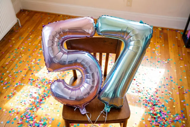 Twee glanzende folieballonnen vormen 57 aan een houten stoel in een zonnige kamer met confetti op de houten vloer en een zilveren en pastelkleurige weerschijn op de ballonnen.