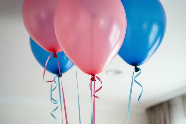 Roze en blauwe ballonnen zweven tegen een wit plafond met lange linten die naar beneden hangen, in zacht licht dat de glanzende ballonnen benadrukt.