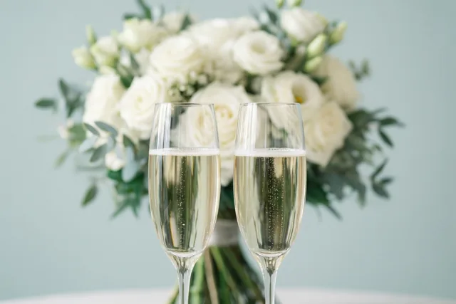 Twee champagneglazen met mousserende wijn staan voor een onscherp wit bruidsboeket, met een lichtblauwe achtergrond en een witte ondergrond.