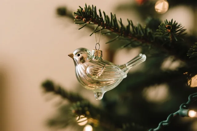 Een doorzichtig glazen vogelornament hangt aan een dennentak van een kerstboom, verlicht door zachte warme lichtjes in groene, gouden en witte tinten.