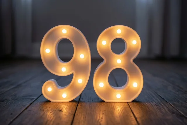 Warm gloeiende lampjes vormen het getal 98 op een donkere houten vloer.