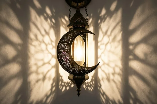 Een messing maansikkellantaarn met fijne uitsparingen hangt tegen een witte muur, met warm licht van binnenuit en een schaduw met patronen op de muur.