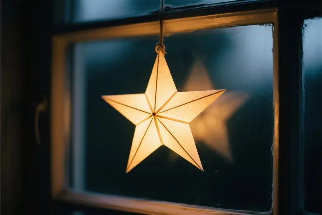 Een lichtgevende papieren ster hangt aan een touwtje voor een donkere ruit, in gouden licht tegen een blauwe achtergrond.