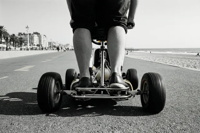 Een zwart-witfoto vanuit een laag standpunt toont de benen van iemand op een gocart op een verharde zeedijk, met rechts strand en zee en gebouwen in de verte.