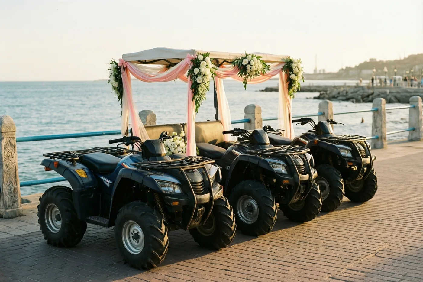 Drie zwarte gocarts staan achter elkaar op een zeedijk bij een stenen balustrade, met roze linten en witte bloemstukken op de voorste en de zee erachter in zacht goudkleurig licht.