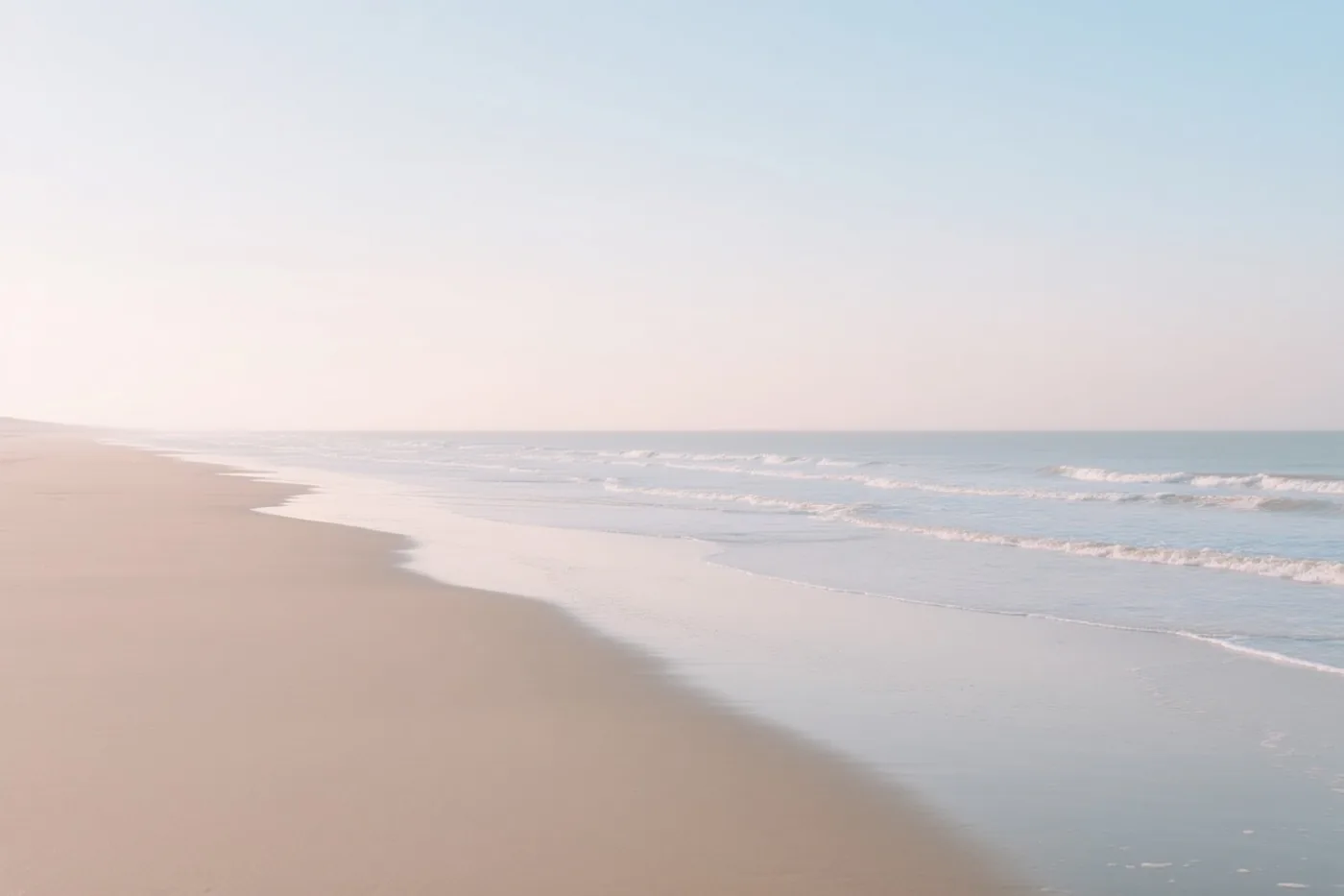 Zachte golven rollen naar het strand onder een bleke lucht in beige, lichtblauwe en lichtroze tinten.