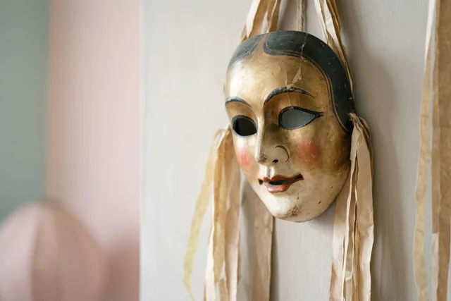 Een oud masker van papier-maché met goudkleurige en zwarte verf en beige papieren slingers hangt aan een muur, met zachtroze en groene tinten op de achtergrond.