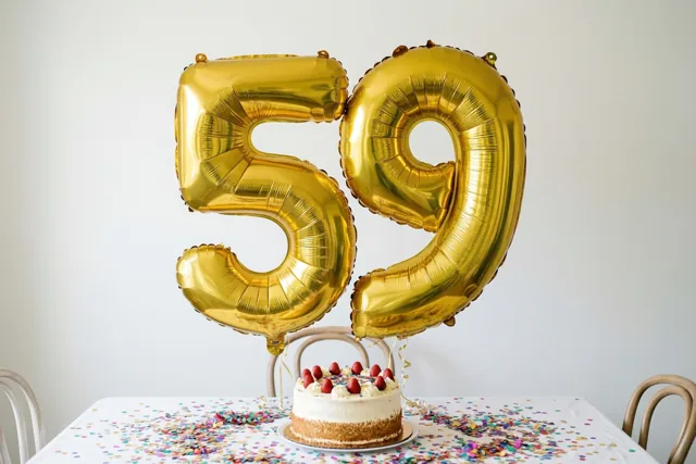 Gouden folieballonnen 59 hangen boven een tafel met kleurrijke confetti en een kleine taart, met stoelen errond tegen een witte muur.