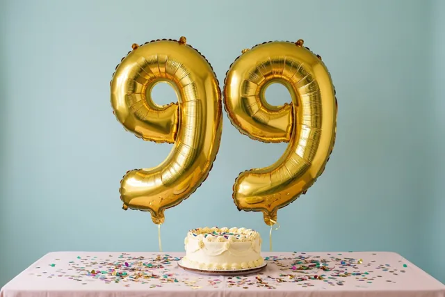 Twee grote gouden folieballonnen vormen 99 boven een roze tafelkleed met confetti en een kleine witte taart, tegen een effen lichtblauwe muur.