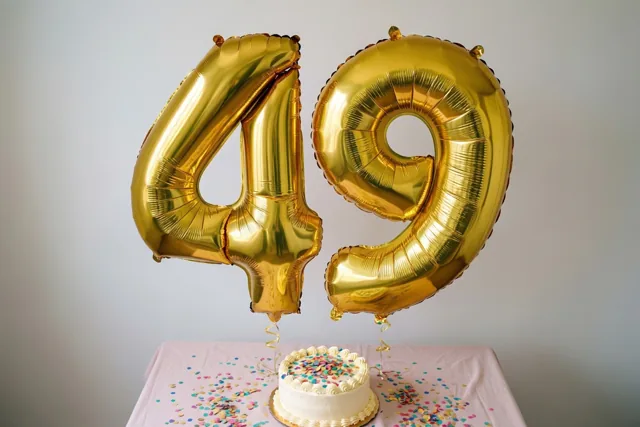 Gouden folieballonnen 4 en 9 zweven boven een tafel met een roze tafelkleed, een witte taart en confetti voor een lichtgrijze muur.