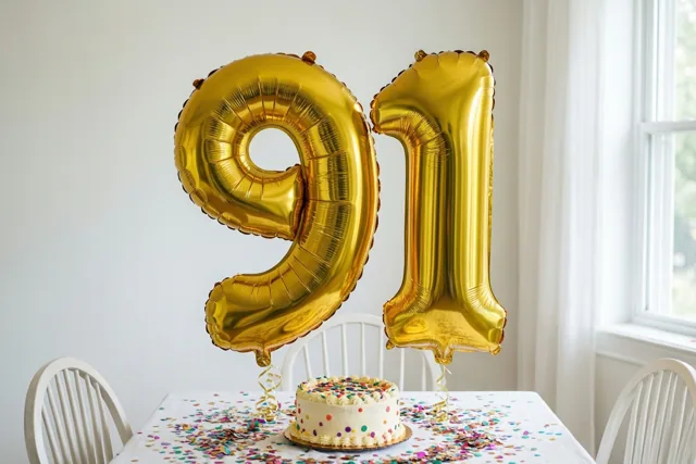 Gouden folieballonnen vormen 91 boven een witte tafel met gekleurde confetti en een kleine taart, bij een raam en witte stoelen.