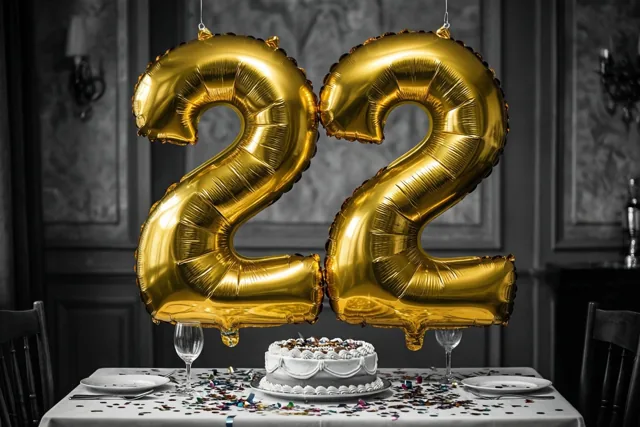 Gouden ballonnen vormen 22 boven een tafel met taart en confetti in een donkere kamer met een zwart-witinterieur.
