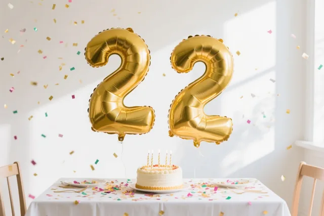 Twee grote glanzende gouden folieballonnen, allebei in de vorm van het cijfer 2, zweven boven een tafel met een taart en confetti in een lichte kamer met witte muren.