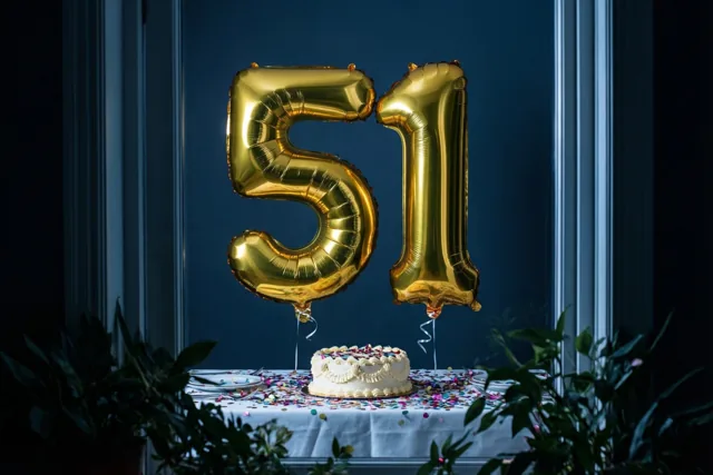 Grote glanzende gouden folieballonnen 5 en 1 zweven boven een tafel met een witte taart en kleurrijke confetti, voor een donkerblauwe muur met groene planten op de voorgrond.