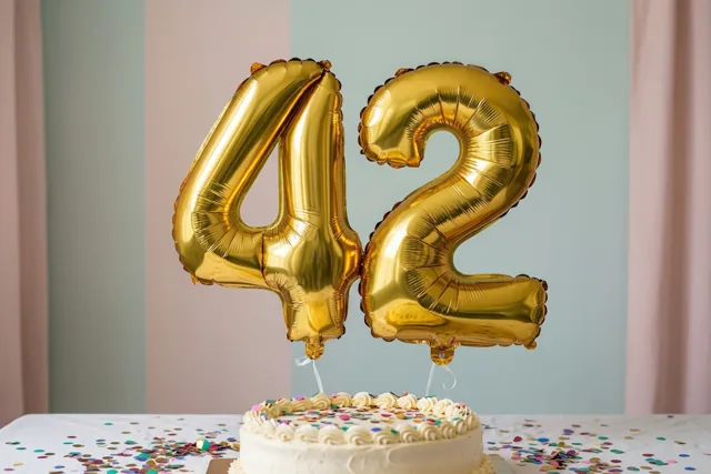 Gouden folieballonnen vormen 42 boven een taart met wit glazuur en kleurrijke confetti op een tafel tegen een gestreepte pastelmuur.