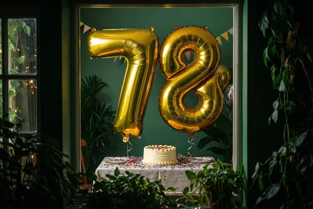 Gouden folieballonnen vormen 78 boven een tafel met confetti en een kleine taart, in een kamer met groene muren, planten, een raam en vlaggetjes.