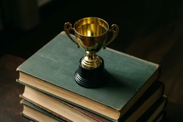 Een kleine gouden beker met twee handvatten staat op oude studieboeken met groene kaften tegen een donkere, onscherpe achtergrond.