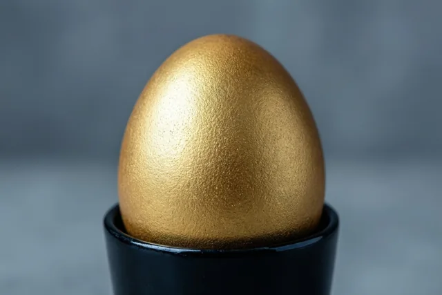 Een goudgeverfd ei staat rechtop in een zwart eierdopje tegen een effen grijze achtergrond.