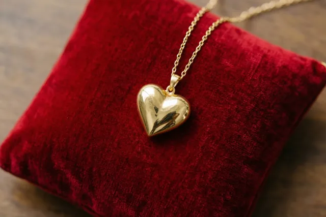 Een gouden hartjeshanger met een fijne ketting ligt op een rood fluwelen kussen op een houten ondergrond in warm licht.