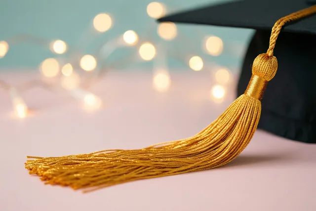 Een gouden kwastje aan een zwarte afstudeerhoed is van dichtbij te zien tegen een zachte pastelkleurige achtergrond met onscherpe warme feestlichtjes.