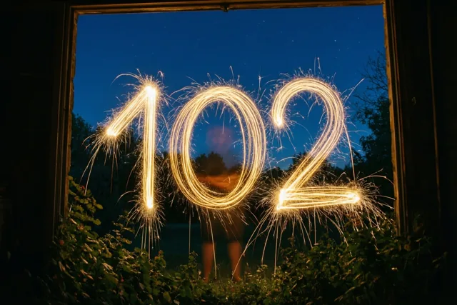 Door een raam zie je gouden lichtsporen van sterretjes die 102 vormen tegen een donkerblauwe nachthemel.