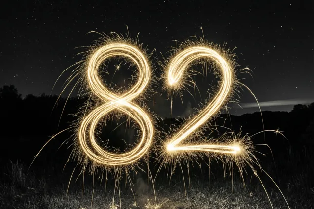 Gouden lichtsporen van sterretjes vormen het getal 82 boven gras, met een sterrenhemel en donkere boomsilhouetten op de achtergrond.