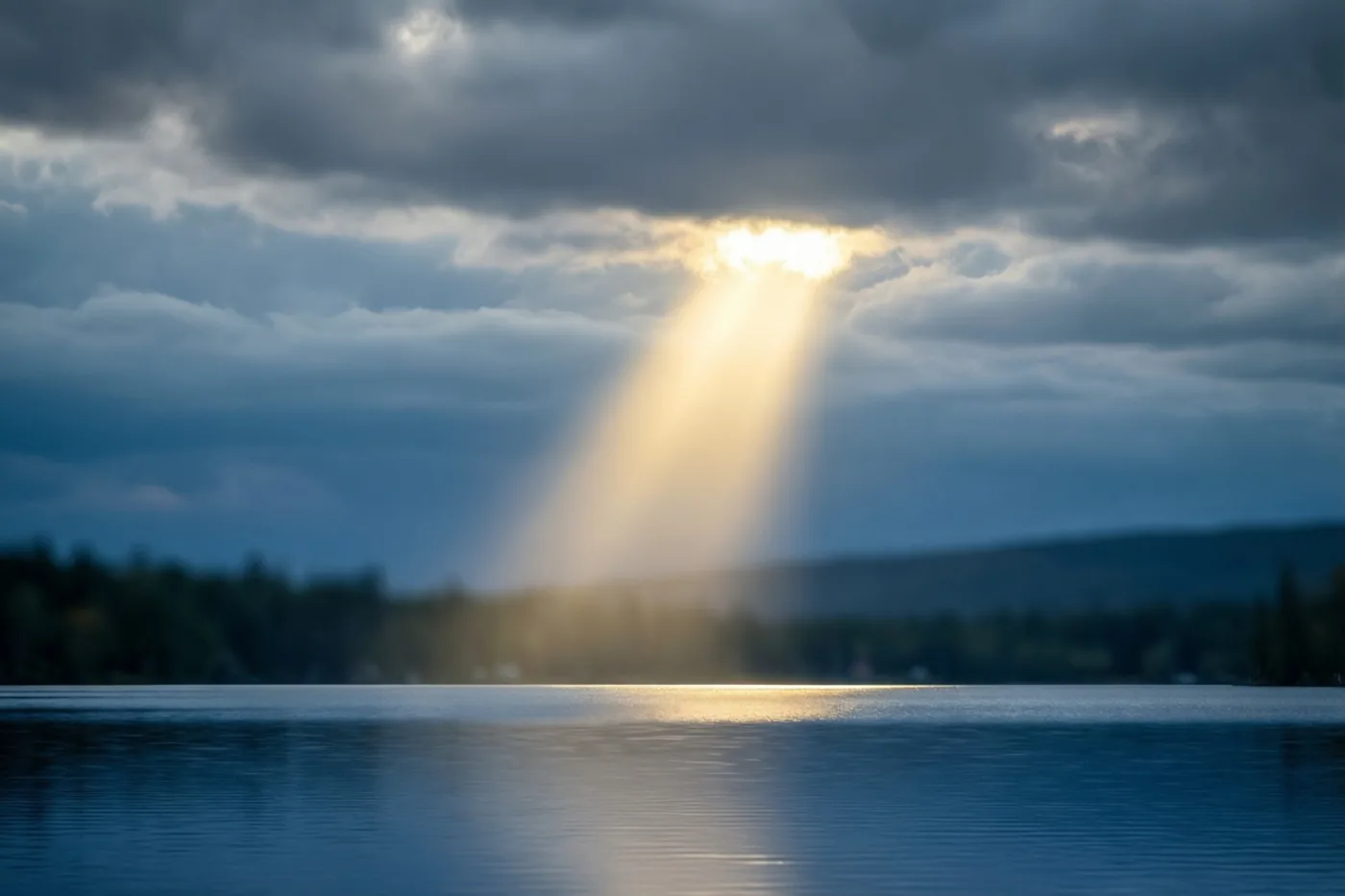 Een gouden lichtstraal breekt door zware grijze wolken en verlicht een kalm meer in blauwe en grijze tinten.