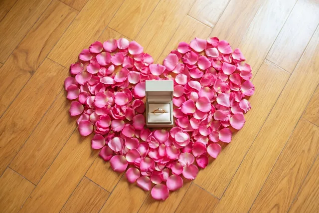 Een hart van roze rozenblaadjes ligt op een lichtbruine houten vloer met in het midden een open lichtgekleurd doosje met een gouden ring.