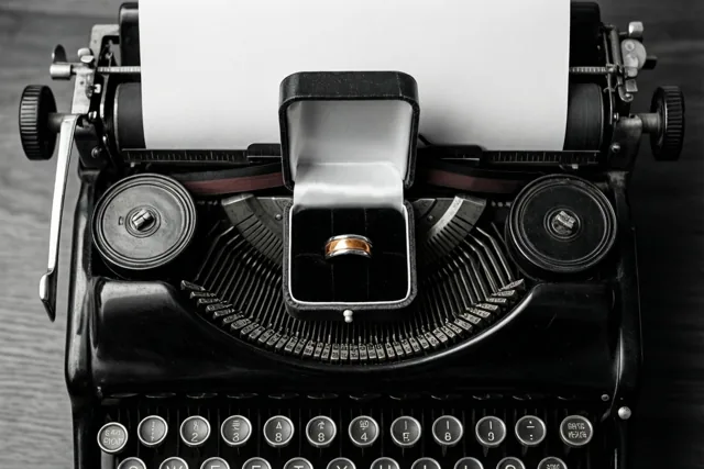 Een open ringdoosje met een gouden ring staat op de toetsen van een vintage typemachine met een blanco vel papier, weergegeven in monochrome tinten.