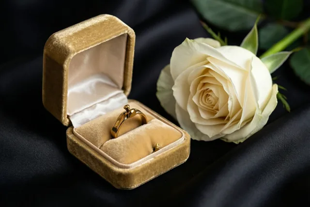 Een open goudkleurig fluwelen ringdoosje met een gouden ring staat naast één witte roos op zwarte zijde in zacht licht.