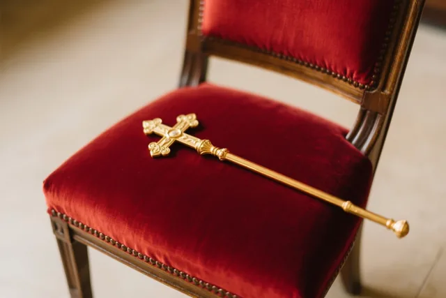 Een gouden staf met een kruisvormige top rust op de zitting van een roodfluwelen stoel met een donker houten frame tegen een lichte, wazige achtergrond.