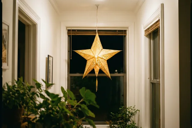 Een vanbinnen verlichte gouden papieren sterlantaarn hangt 's nachts aan het plafond van een witte gang met potplanten op de voorgrond en ingelijste afbeeldingen aan de muren.