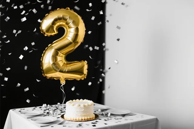 Een grote gouden cijferballon 2 zweeft boven een tafel met een wit geglazuurde taart en zilveren confetti, tegen een zwart-witte achtergrond.