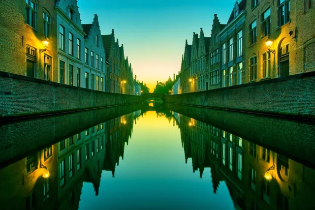 Een kanaal in een oud Vlaams stadje ligt bij zonsopgang tussen historische huizen met puntdaken en warme straatlampen, met goudgele weerspiegelingen in blauwgroen water.
