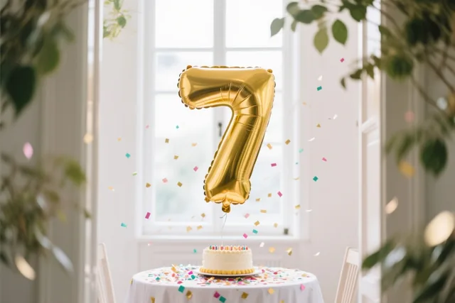 Een grote gouden folieballon in de vorm van een 7 zweeft boven een tafel met een wit kleed, een kleine taart en kleurrijke confetti, bij een licht raam met groen rondom.