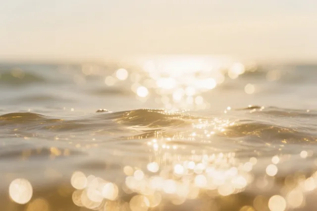 Zonlicht schittert op zachte zeegolven in goudkleurige, beige en witte tinten.